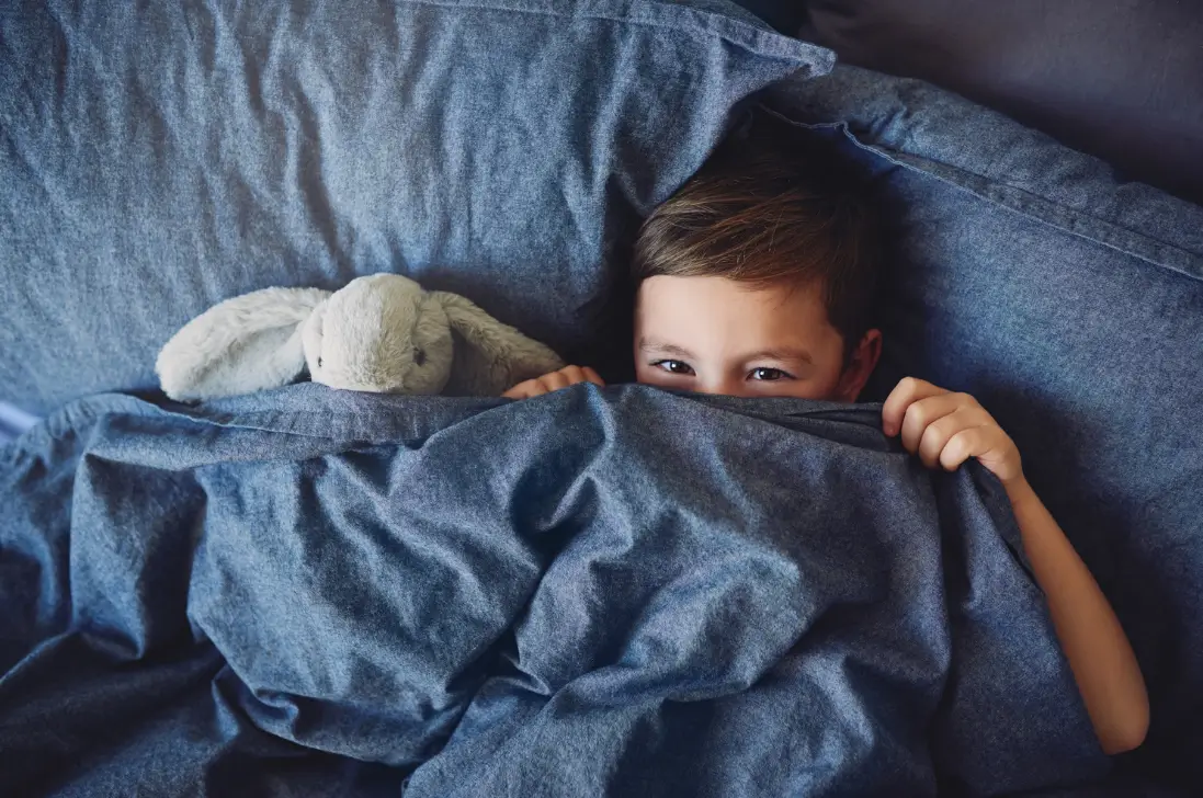 bedwetting-alarms-do-they-really-work Young child peeking over a blue duvet.