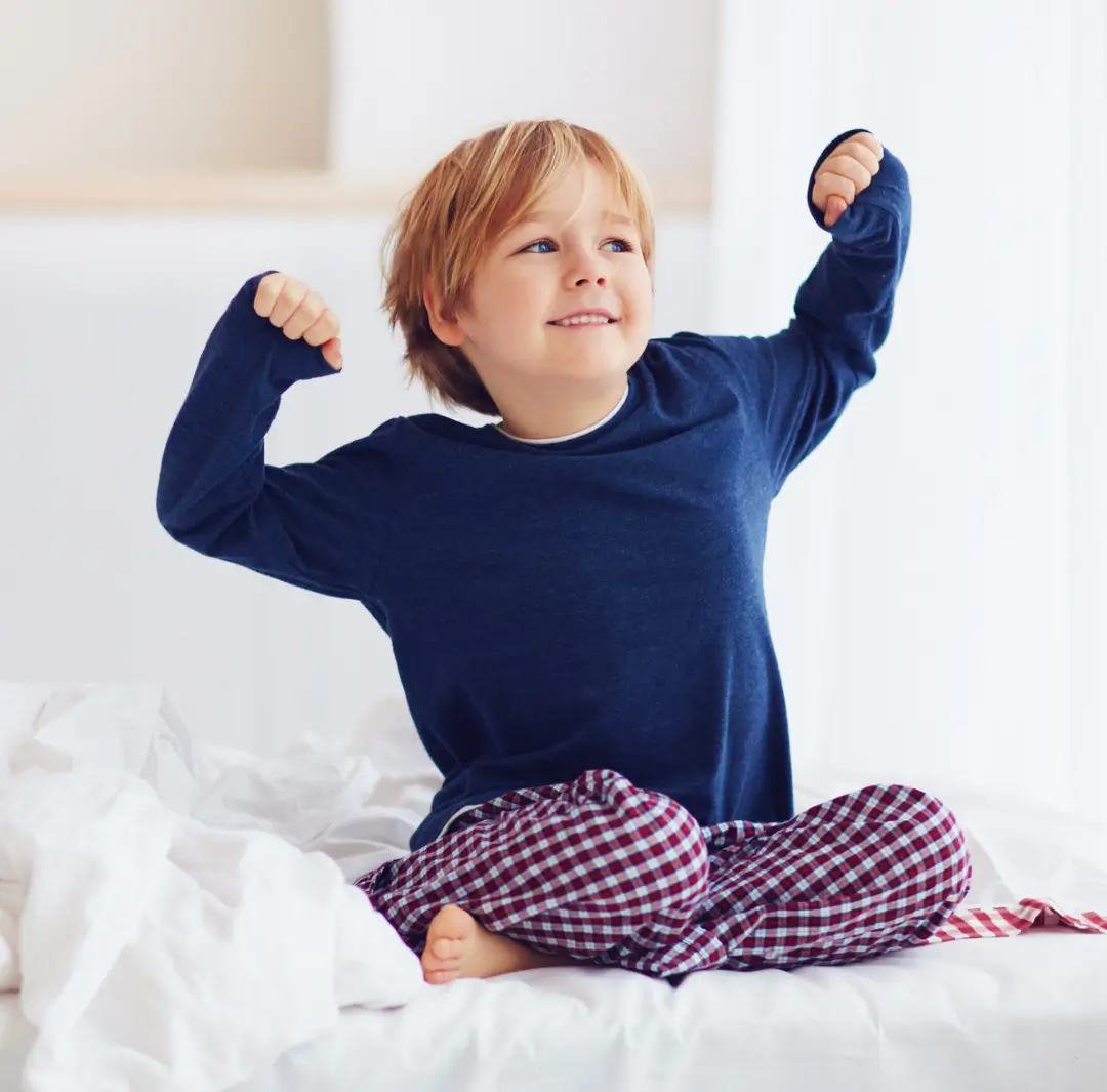 Smiling child sitting on bed and flexing arms proudly after overcoming bedwetting with a natural bed wetting hypnosis program.